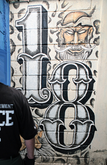 A large painted '18' with a bearded face on a cinder-block wall, an officer standing in the foreground